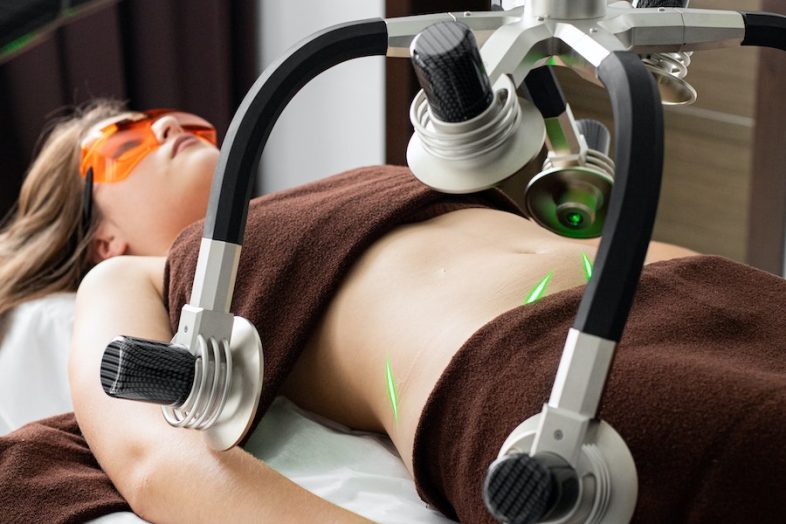 Female client in eyewear lying on table under modern laser fat loss machine during liposuction treatment in light medical clinic