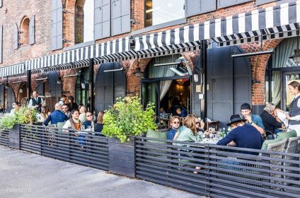 People enjoying outdoor dining at a stylish cafe with brick facade and striped awnings in a lively urban setting