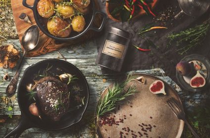 Rustic dining scene with roasted potatoes, steak in skillet, figs, herbs, and spices on a wooden table setting