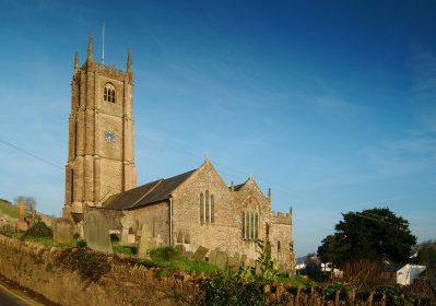 Medieval stone church with tall clock tower in rural England, surrounded by gravestones and greenery under a clear blue sky