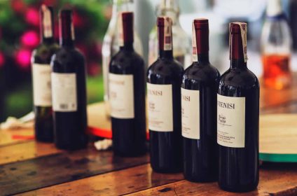 Row of red wine bottles on a rustic wooden table, with blurred background of greenery and flowers