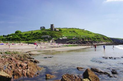 Sandy beach with people and rocky shore, green hill and historic building under clear blue sky in Cornwall, England