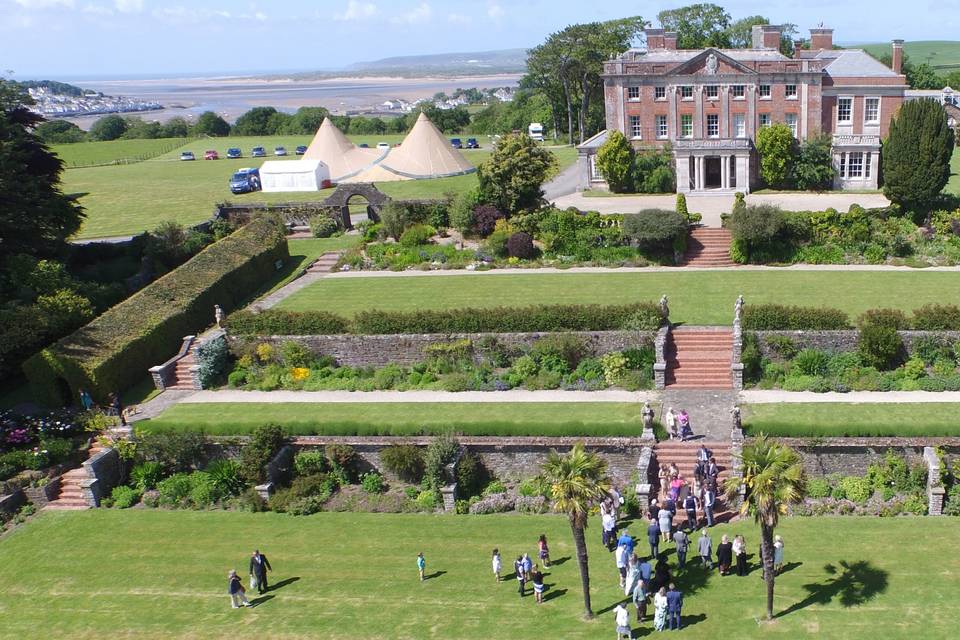 Aerial view of historic mansion with landscaped gardens and guests, overlooking countryside and sea on a sunny day