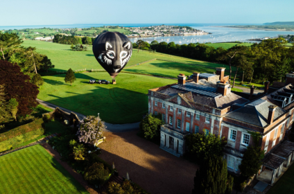 Aerial view of a historic manor and fields with a wolf-themed hot air balloon near the coast in Devon, England