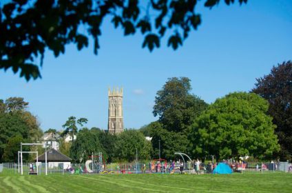 Playground and church tower under blue sky, surrounded by lush greenery in a scenic park setting