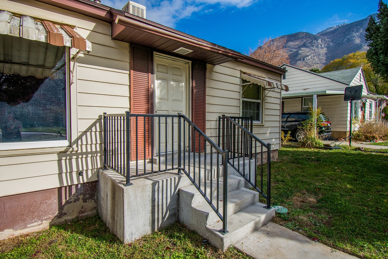 Front steps of a small house with metal railing on a sunny day, surrounded by greenery and mountain view