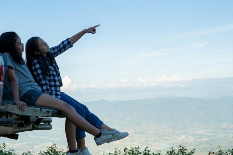 Two girls sitting on a truck enjoying a scenic mountain view, one pointing at the sky