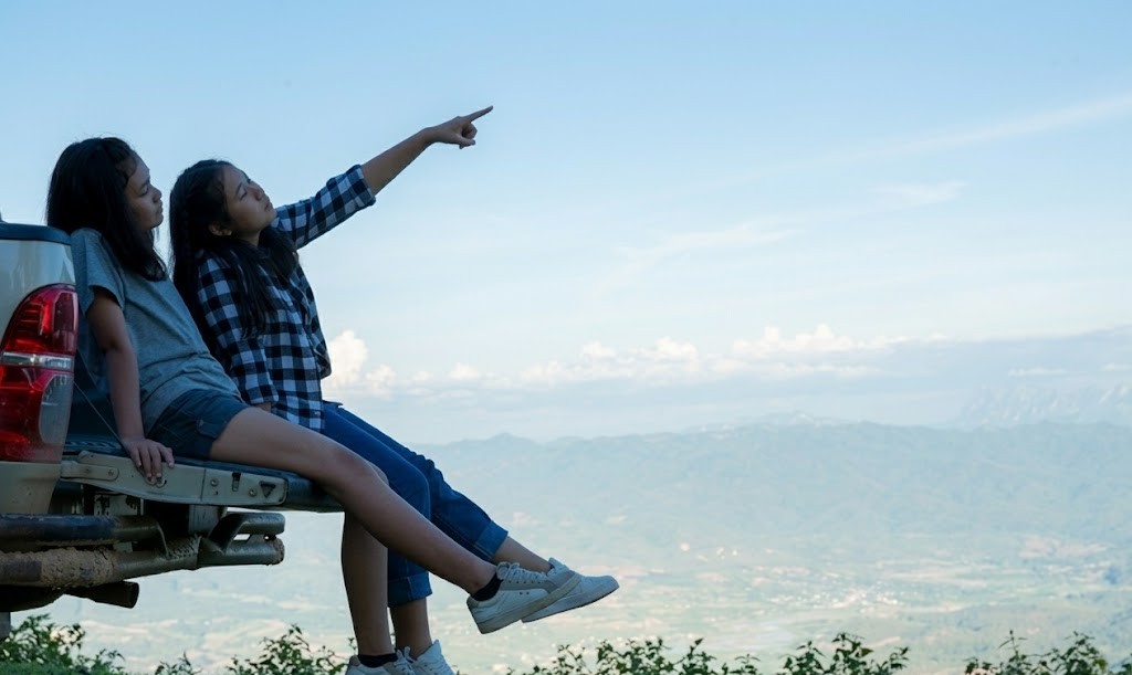 Two girls sitting on a truck enjoying a scenic mountain view, one pointing at the sky