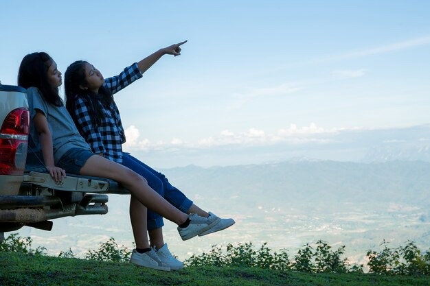 Two young women sit on a truck's back, pointing excitedly at something in the distance