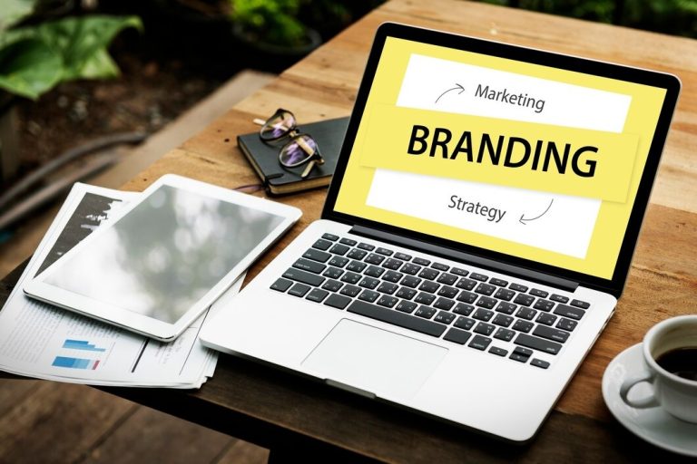 Branding design displayed on a laptop beside a cup of coffee and a stack of papers on a wooden table.